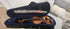 A violin and bow in a blue-lined black case resting on a light-colored couch.