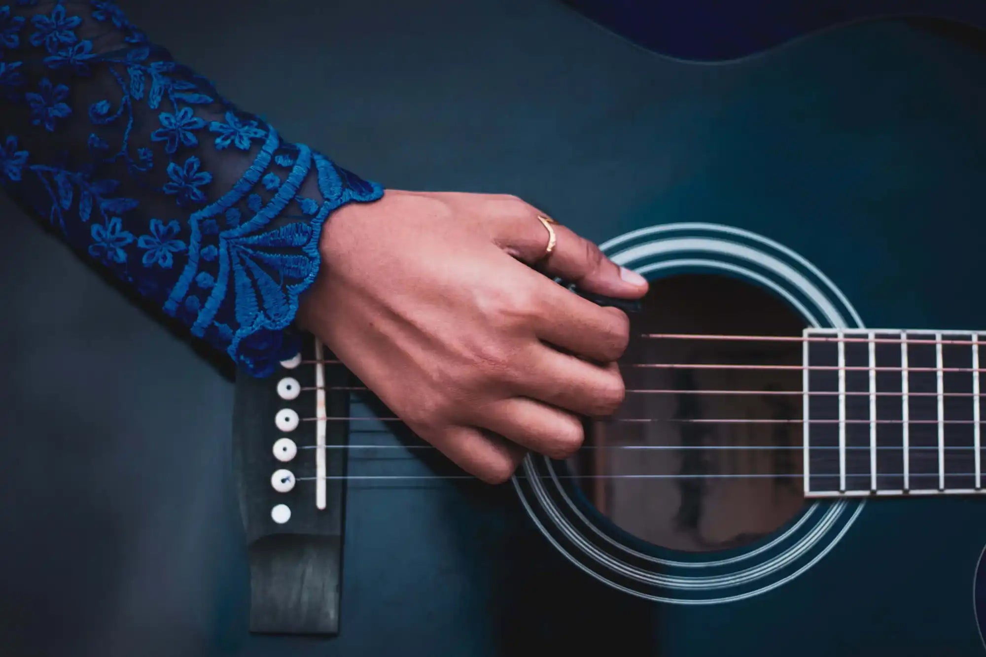 A hand wearing a ring is strumming the strings of a black acoustic guitar. The person is wearing a blue, floral-patterned sleeve.