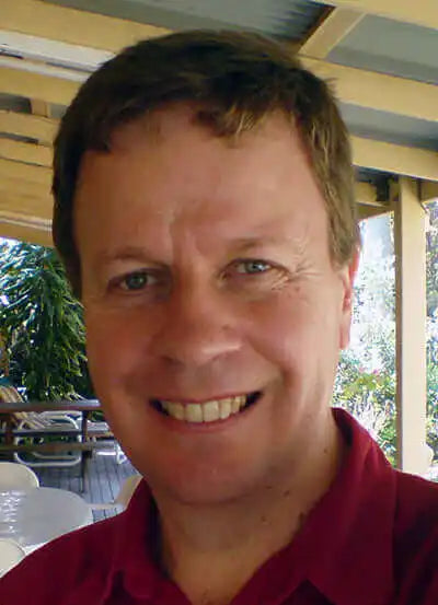 A man with short brown hair wearing a red shirt smiles at the camera under a covered outdoor patio.