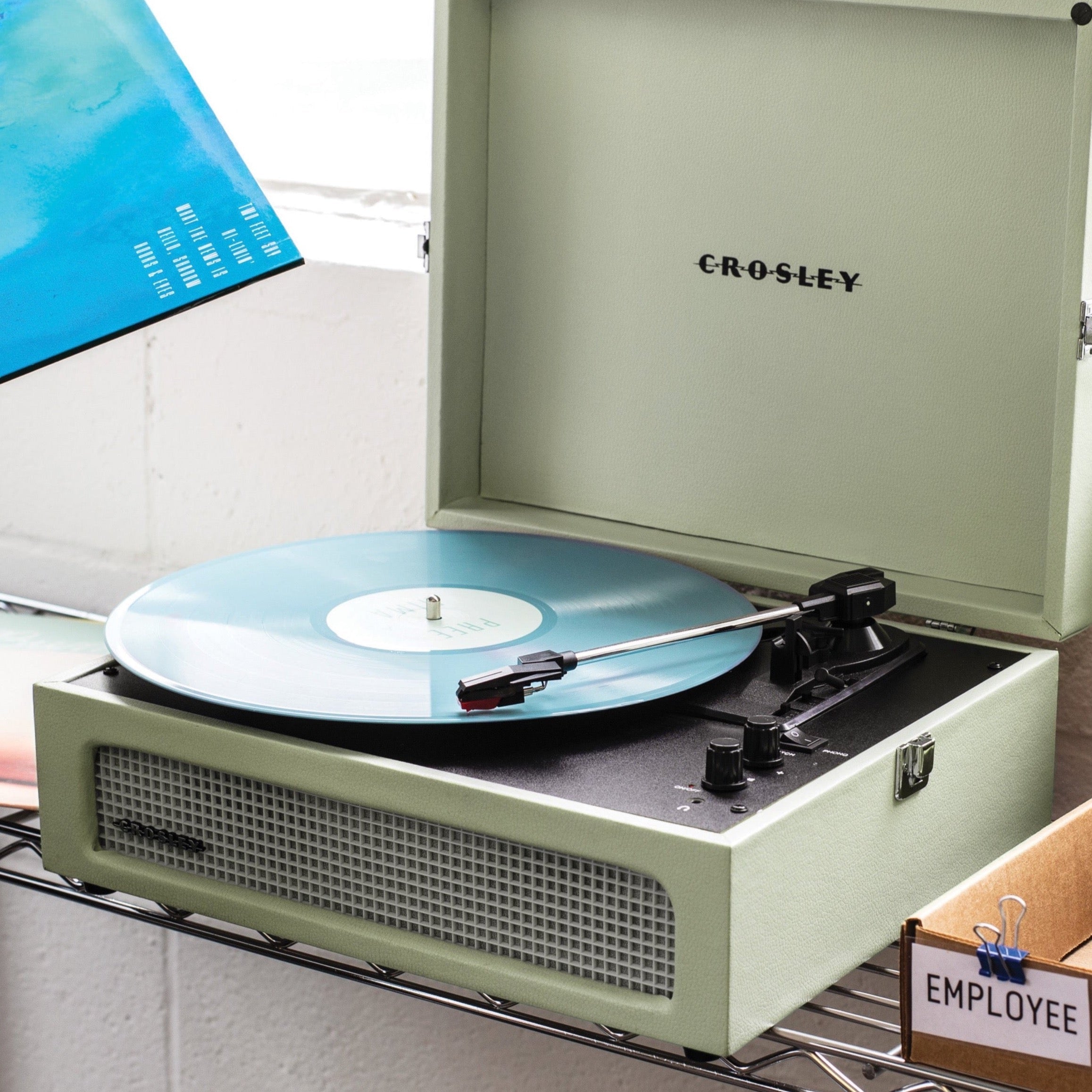 The Crosley Voyager Sage Bluetooth Portable Turntable by Crosley is shown with the lid open, playing a vinyl record. The front panel includes a speaker grille, control knobs, and RCA outputs for easy connectivity.