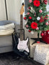 A black electric guitar on a stand next to a Christmas tree decorated with red and white ornaments, a gift bag, and a grey armchair with cushions.