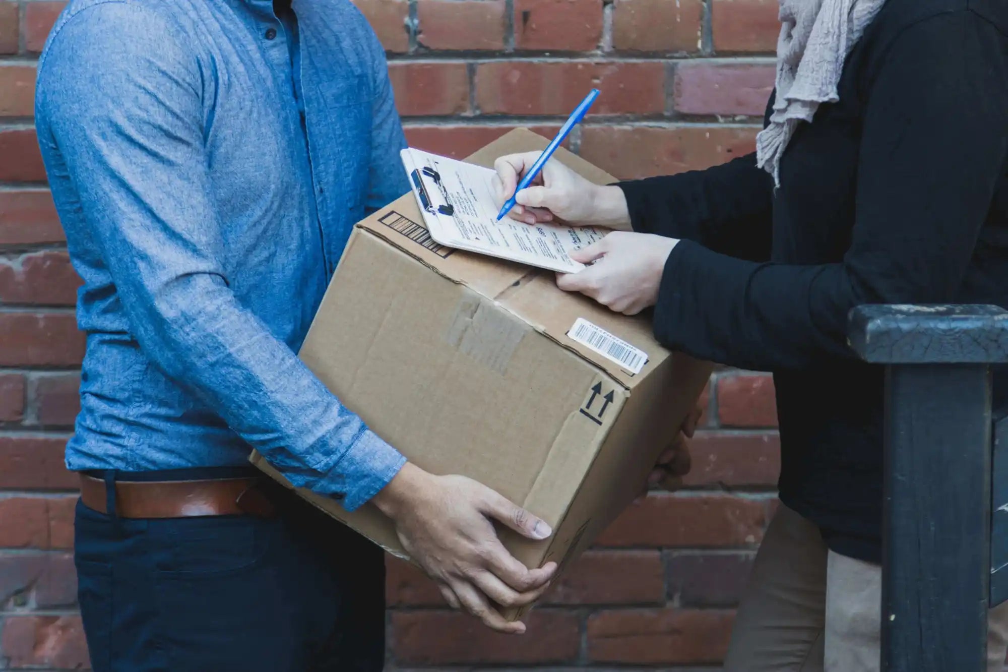 Two people stand by a brick wall; one hands over a cardboard box while the other signs a delivery receipt on a clipboard.