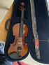 A violin in a hard case with a bow stored in a protective plastic sleeve next to it, placed on a wooden table.