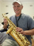 A man wearing a white cap and gray shirt is smiling while holding a gold alto saxophone indoors.