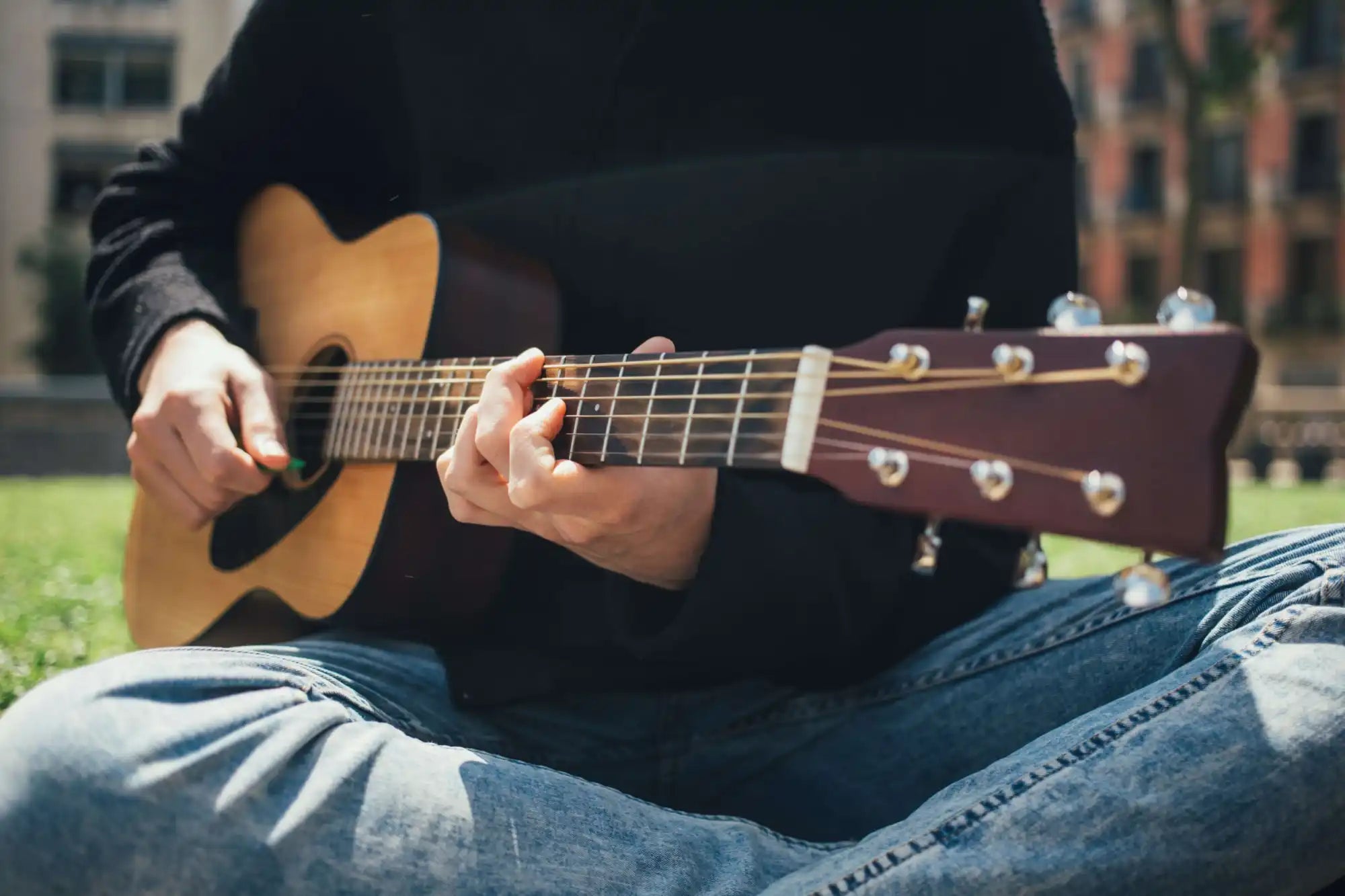 Person sitting cross-legged outdoors, wearing jeans and a dark top, playing an acoustic guitar. The photo focuses on the guitar and hands.