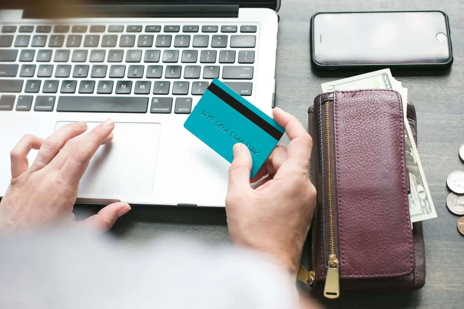 Person holding a credit card and using a laptop, with a wallet, cash, coins, and a smartphone on the table nearby.