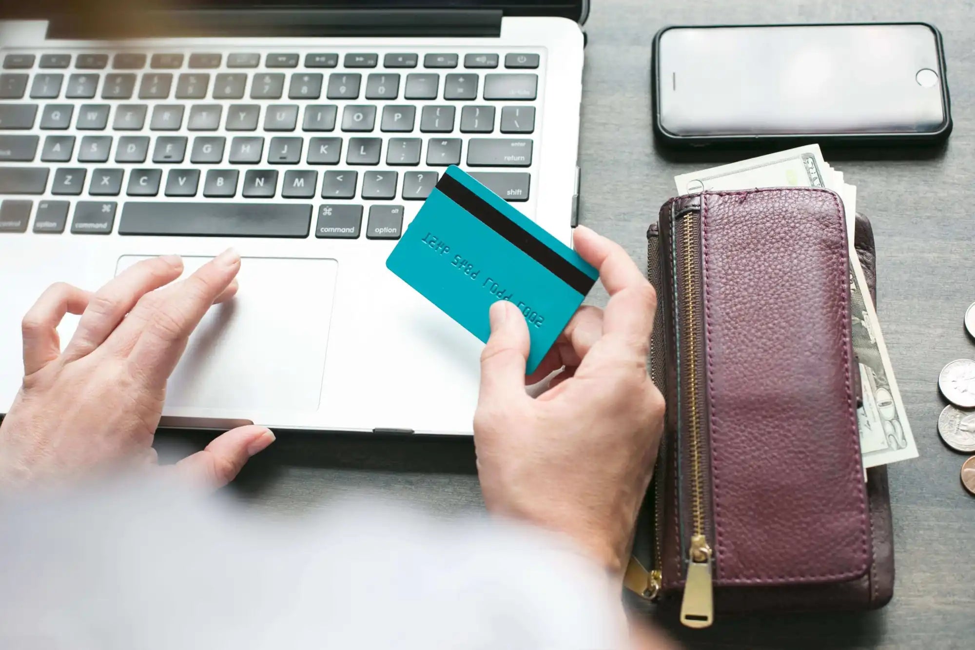 Person holding a credit card and using a laptop, with a wallet, cash, coins, and a smartphone on the table nearby.
