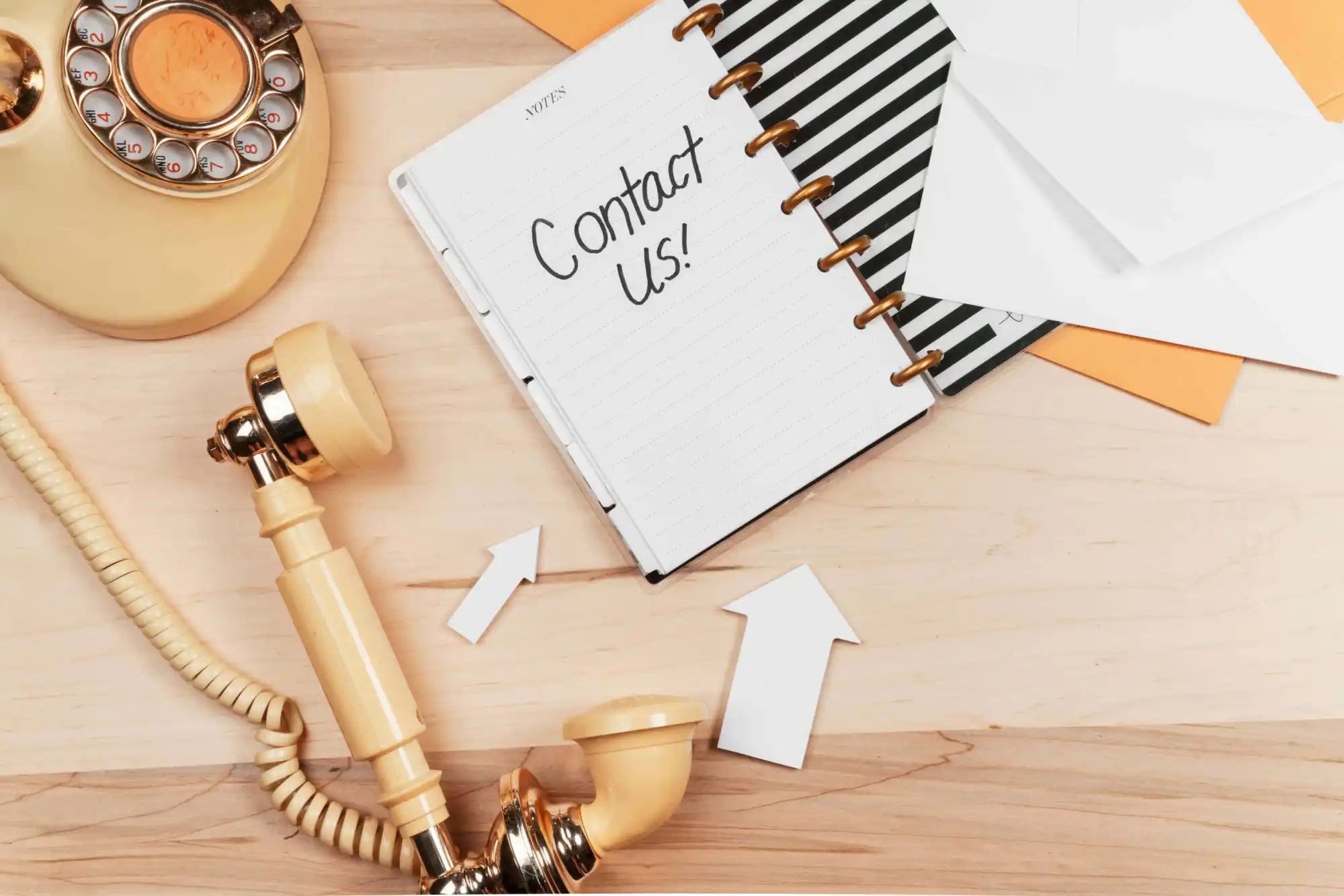 A vintage rotary phone, an open notebook with Contact Us! written inside, white envelopes, and paper arrows arranged on a wooden desk.