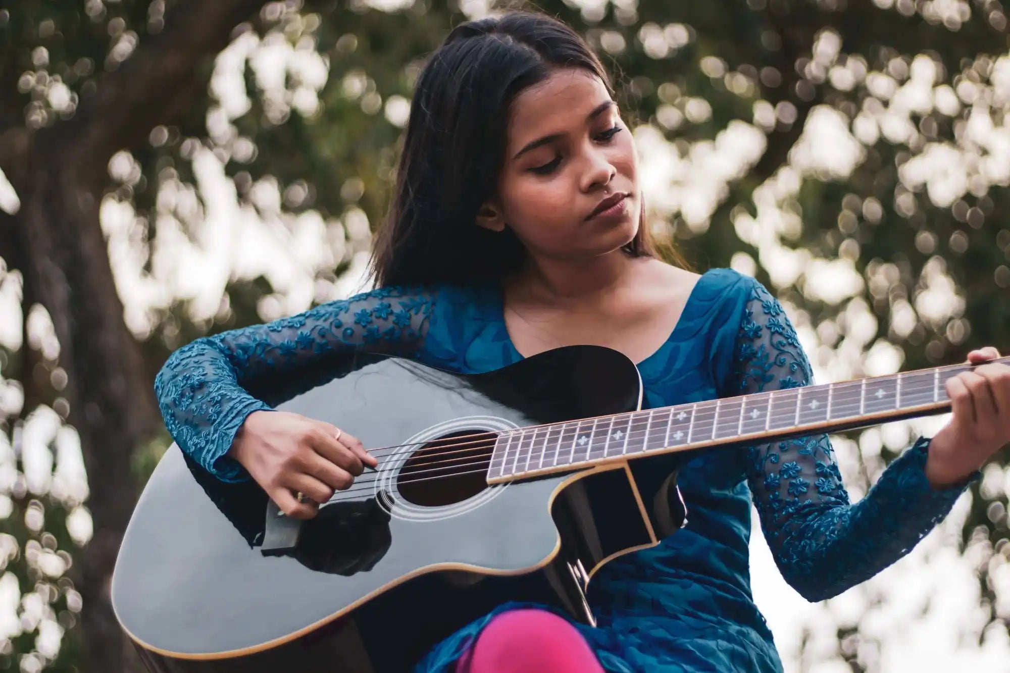 A young girl in a blue dress plays a black acoustic guitar outdoors, with trees blurred in the background.