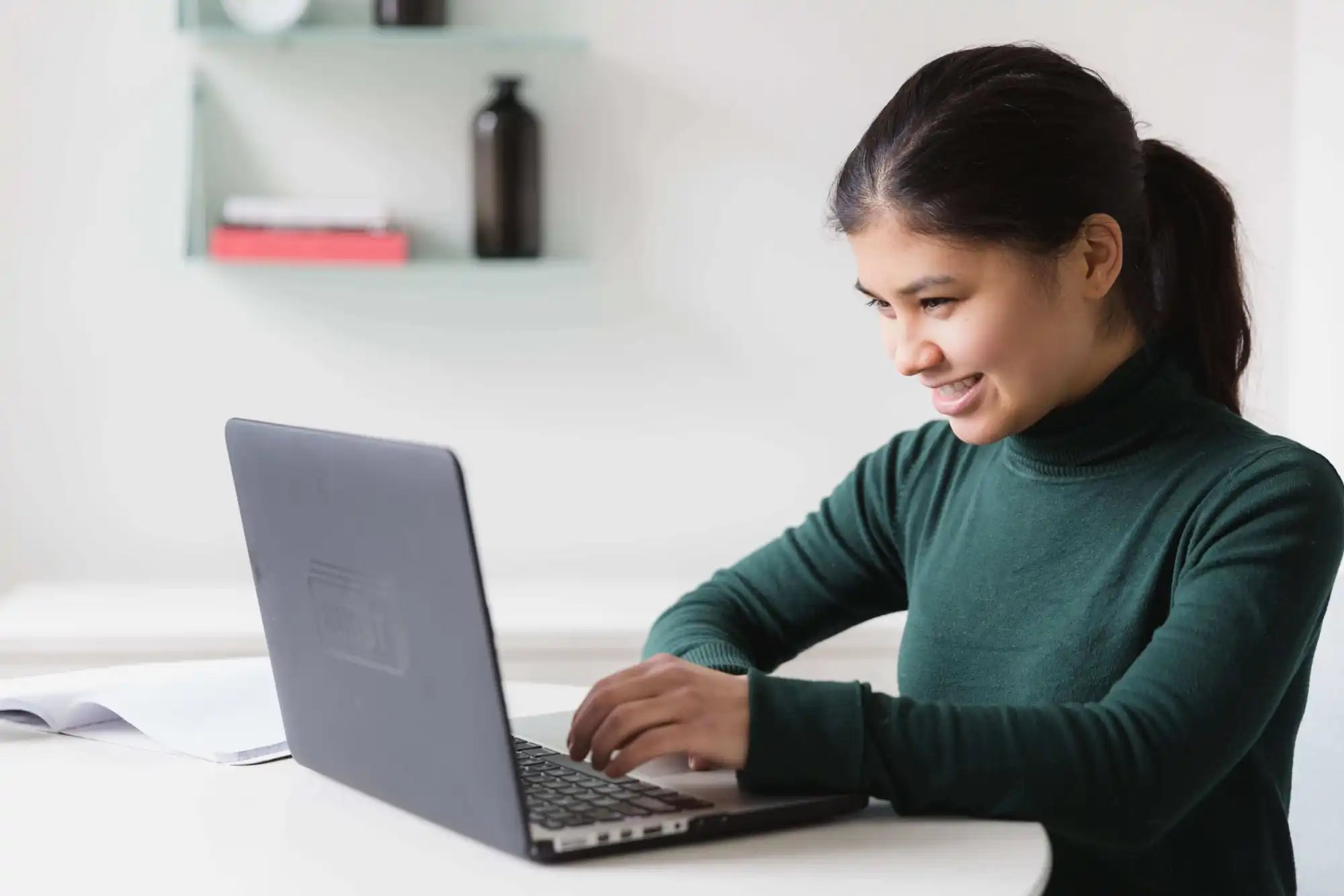 A woman in a green turtleneck sits at a desk, typing on a laptop and smiling, with an open notebook beside her. Shelves with books and a vase are in the background.